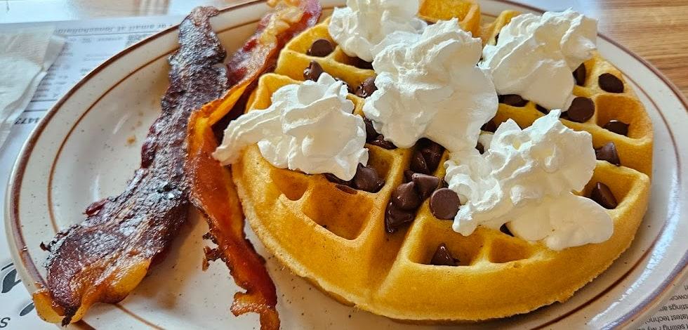 A Belgian waffle with chocolate chips and whipped cream, with two strips of bacon.