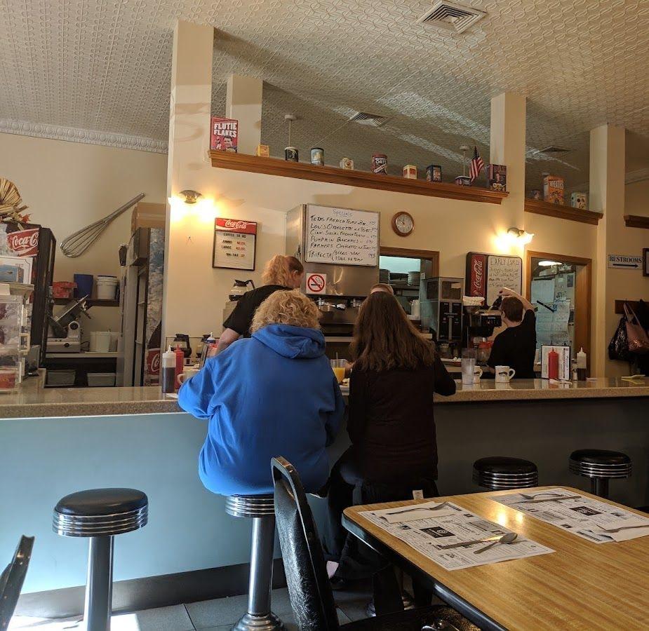 Customers at the counter, with the day’s specials written on a whiteboard behind it and cereal boxes lined up on the shelf above.