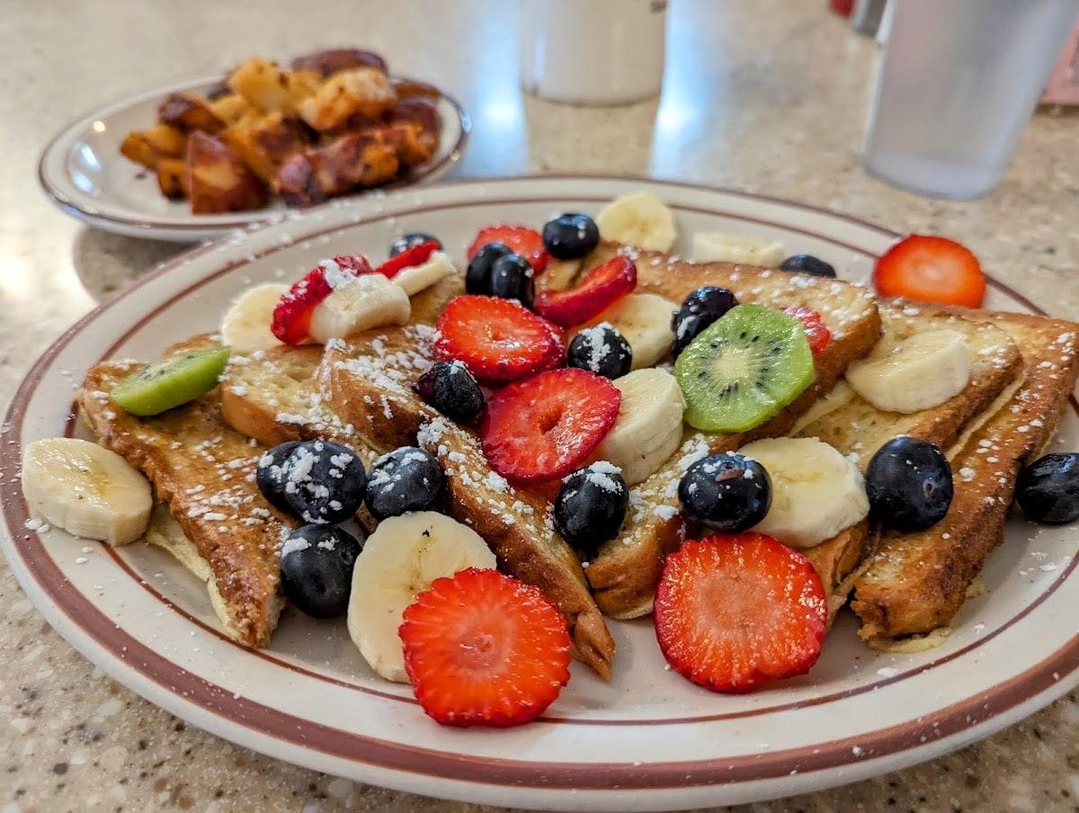 Fruitopia on the counter: French toast dusted with powdered sugar under sliced strawberries, bananas, blueberries and kiwi, with home fries behind.