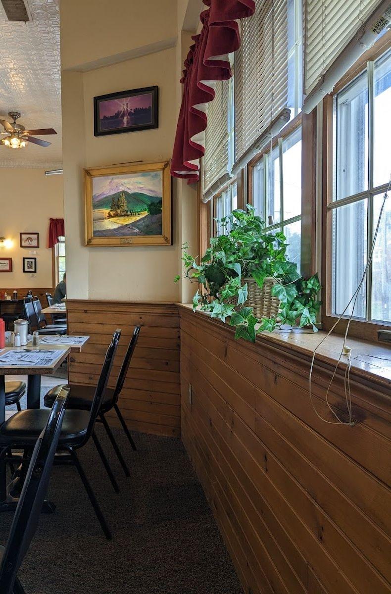 A corner of the dining room: wood paneling, framed paintings, an ivy plant on the windowsill and red valances over the windows.